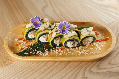 Taze suşi ruloları, yenilebilir çiçekler ve dilimlenmiş bademlerle süslenmiş ahşap bir tabakta servis edilir. Ayarlar yemeğin canlı renklerini ve dokularını vurgular.