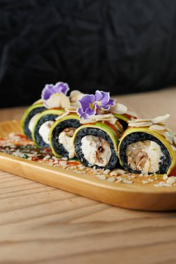 Suşi ruloları ahşap bir tabakta güzelce düzenlenir. İçinde çiçekler ve fındıkla süslenmiş avokado ve balık gibi malzemeler var. Lezzetli bir Japon yemeği..