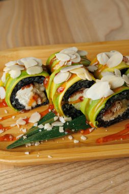 Yengeçle doldurulmuş suşi ruloları ve avokado dilimlerine sarılmış ahşap bir tabakta oturuyorlar. Badem dilimleri ve sos ekmeklerin etrafına serpilmiş. Taze yeşil yapraklar renk ekler.