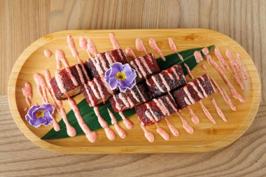 Çeşitli yiyecek ürünleri, ahşap bir tabakta düzgün bir şekilde düzenlenir. Yemekler renkli katmanlar ve soslar içeriyor. Bir çiçek kuruluma görsel ilgi ekler.