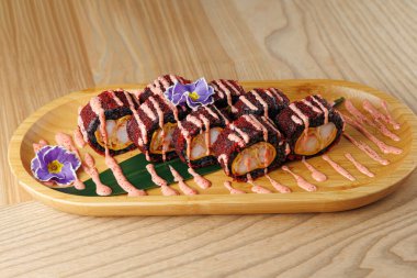 Taze suşi ruloları ahşap bir tabakta, sosla kaplı ve yenilebilir çiçeklerle süslenmiş. Bu yemek Japon mutfağında tipik bir tat ve renk karışımı sergiliyor..