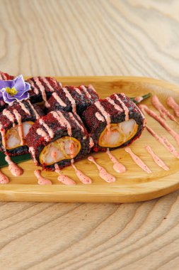 Suşi ruloları üzerine renkli soslar serpiştirilmiş ahşap bir tabakta düzenlenir. Tabakta karides ve dekoratif bir çiçek var. Bu yemek geleneksel Japon lezzetlerini sergiliyor..
