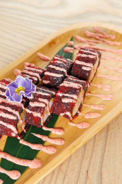 Suşi dürümleri ahşap bir tepside düzgünce düzenlenir. Üstünde dekoratif bir çiçek ve etrafa serpiştirilmiş soslar vardır. Parlak renkler yemeğin görünümünü güçlendirir.