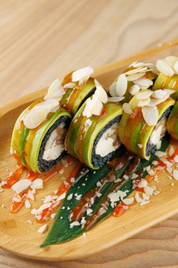 Avokado, pilav ve tavukla yapılan suşi ruloları ahşap bir tabakta güzelce düzenlenir. Dilimlenmiş badem ve sos ekmeklerin etrafına saçılmış. Bu yemek canlı tatlar sunuyor..