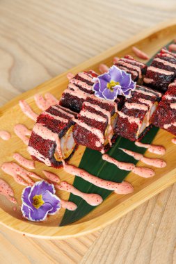 Suşi hamuru ahşap bir tabakta rengarenk bir sos ve yenilebilir çiçeklerle süslenmiş. Sunum yemeğin ince detaylarını vurgular.