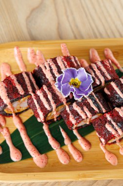Suşi ruloları ahşap bir tepsiye güzelce yerleştirilir, pembe sos ve mor bir çiçekle süslenir. Tabakta Japon mutfağını vurgulayan çeşitli doku ve renkler yer alıyor..
