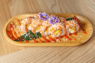 Suşi ruloları ahşap bir tabakta güzelce düzenlenir. Dekoratif çiçekler üstte durur, soslar da ustalıkla etrafa serpilmiştir. Masası ahşap olan bir restoran..