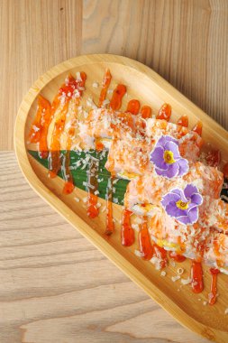 Suşi ruloları, yenilebilir çiçekler ve sos çizgileriyle süslenmiş ahşap bir tabakta güzelce düzenlenir. Tabak, bir restorandaki ahşap bir masada servis edilir..