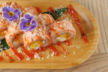 Pirinç ve sebzelerle dolu suşi ruloları ahşap bir tabakta düzenlenir. Tabak somon, renkli soslar ve mor çiçeklerle kaplı..