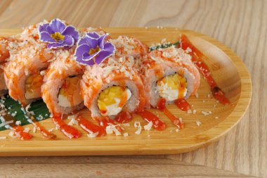 Suşi ruloları ahşap bir tabakta güzelce düzenlenir. Ekmekler somon, yumurta, sos ve yenilebilir çiçeklerle süslenmiş. Tabaklar basit bir ahşap masaya yerleştirilmiş..