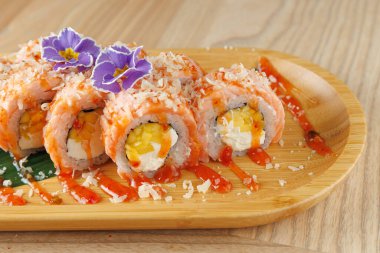 Renkli suşi ruloları ahşap bir tabakta oturur. Ekmeklerde yumurta ve peynir dolgusu, yengeçle kaplanmış ve sunum için çiçekler ve sosla süslenmiş..