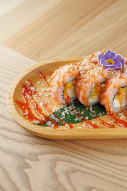 Suşi ruloları ahşap bir tabağa konur, sos ve çiçekle süslenir. Renkli malzemeler masanın üzerinde lezzet ve dokuların bir karışımını sergiler..