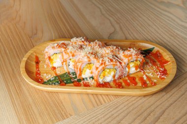 Suşi tabağında karidesle kaplanmış ve sosla süslenmiş birkaç rulo var. Rahat bir yemek alanında, ahşap bir tepside servis ediliyor..