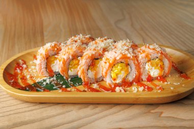 Suşi ruloları taze balık, pirinç ve dolgular ahşap bir tabakta sunulur. Renkli yemek, canlı malzemeler ve Japon mutfağını ön plana çıkaran sos içeriyor..