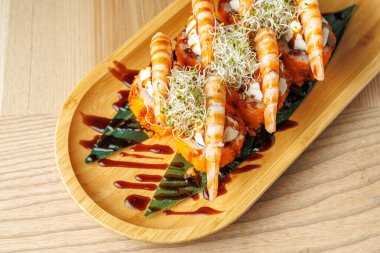 Karides ve suşi ruloları üzerine sos serpiştirilmiş ahşap bir tepside düzenlenir. Taze malzemeler lezzet ve dekorasyon için eklenir. Güzel bir yemek deneyimi belli oluyor..