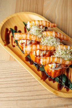Karidesli suşi ruloları ahşap bir tabakta oturur. Sosla kaplanmış ve lahanayla süslenmiş. Sahne geleneksel Japon mutfak sanatı ve sunumunu vurguluyor.