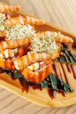 Karidesli suşi ruloları ahşap bir tabakta düzenlenir. Her rulo karides ve lahanayla kaplıdır, üzerine sos serpilmiştir. Bu yemek çeşitli mutfaklardan çeşitli tatlar içeriyor..