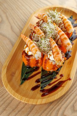 Çeşitli suşi dürümleri ahşap bir tabakta oturur. Her rulo karidesle kaplanmış ve lahanayla süslenmiş. Bir tutam sos tabağa tat katar..