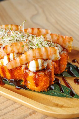 Tahta bir tabakta karides ve sosla süslenmiş suşi ruloları sergilenir. Bu yemek çeşitli tatları ön plana çıkarıyor, Japon mutfak sanatını bir yemek deneyiminde sergiliyor..