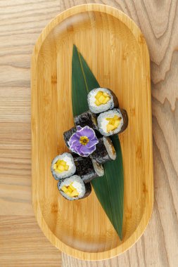 Taze mangolarla dolu beş suşi rulosu bir yaprak üzerinde düzenlenir ve ahşap bir tabakta sunulur. Yenilebilir bir çiçek yemeğe biraz renk katar..