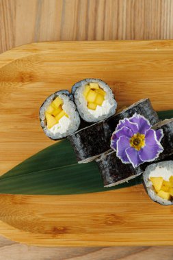 Mango parçalarıyla dolu suşi ruloları bambu bir tahtanın üzerinde duruyor. Mor bir çiçek dekoratif bir dokunuş ekler. Bu yemekte taze malzemeler ve ustaca hazırlanmış yemekler var..