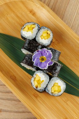 Mango ve krem peynirle dolu suşi ruloları ahşap bir tabakta ustalıkla düzenlenir. Mor bir çiçek biraz renk katar. Bu yemek lezzet ve dokuların bir karışımını sergiler..