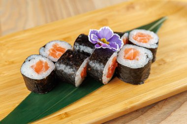 Somon içeren suşi ruloları ahşap bir tabakta güzelce düzenlenir. Mor bir çiçek süsleme ekler. Bu ortam bir restoranda sıradan bir yemektir..