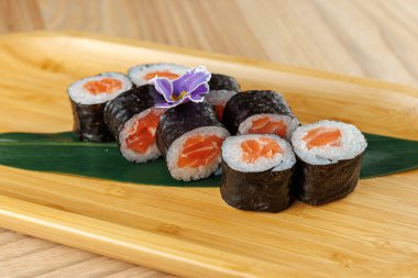 Suşi ruloları ahşap servis tabağına düzgünce yerleştirilir ve deniz yosununa sarılmış taze somon parçaları sergilenir. Küçük bir çiçek sunuma biraz stil katar.