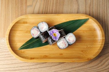 Çeşitli suşi dürümleri ahşap bambu tabakta sunuluyor. Suşi canlı renklere ve yemeği süsleyen narin bir çiçeğe sahiptir..
