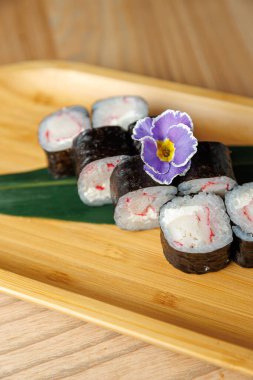 Pirinç ve yengeçle yapılan suşi ruloları bambu tabakta güzelce düzenlenir. Mor, yenilebilir bir çiçek suşinin yanına renk katar ve büyüleyici bir sunum yaratır..