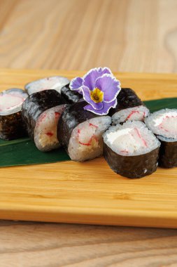 Taze suşi ruloları ahşap bir tahtada renkli bir çiçek süslemesi ile düzenlenir. Ekmekte deniz yosunu, pirinç ve dolgu var. Canlı bir yemek seçeneği yaratıyor..