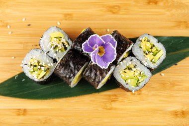 Avokado dolu renkli suşi ruloları ve bambu kesme tahtasına serpiştirilmiş susam tohumları. Çiçek süslemesi yemeğe biraz detay katar..