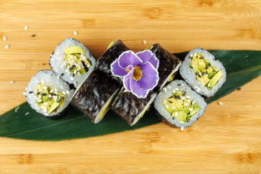 Avokado, salatalık ve pirinçle dolu suşi ruloları ahşap bir tahtada güzelce düzenlenir. Renkli bir çiçek tabağa dekoratif bir dokunuş ekler. Bu Japon mutfağının cazibesini gösteriyor..
