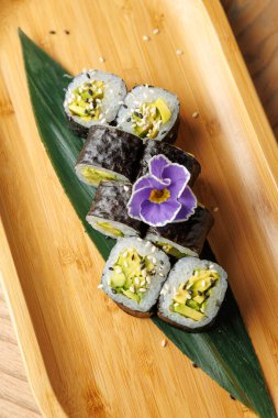 Suşi rulosu koleksiyonu ahşap bir tepside duruyor. Ekmeğe taze sebze konur ve renkli bir çiçekle süslenir. Bu güzel yemeğin tadını çıkarın..