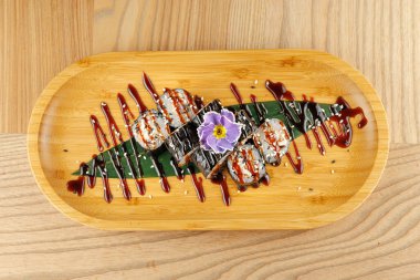 Suşi ruloları ahşap bir tabakta serpiştirilmiş sos ve bir çiçek süslemesi ile düzenlenir. Huzurlu bir yemek düzeni bu yemeğin görsel çekiciliğini arttırır..