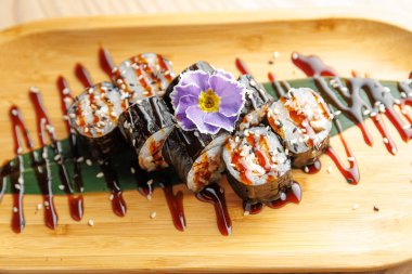 Suşi ruloları üzerinde çiçek olan ahşap bir tabakta düzenlenir. Ekmeğe sos serpilmiş ve susam tohumları serpiştirilmiş. Asya mutfağı sanatı sergileniyor..