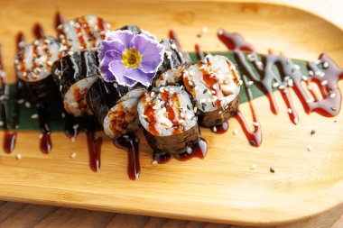 Suşi ruloları bambu tabakta güzelce düzenlenir. Yemek, ekmeklerin üzerine serpiştirilmiş sos ve garnitür olarak bir çiçek içeriyor. Çekici bir sunum oluşturuyor..