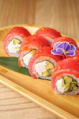 Krem peynir ve avokado ile dolu suşi ruloları ahşap bir tabakta düzenlenir. Mor bir çiçek biraz renk katar. Sahne, suşinin tadını çıkardığı bir yemek sahnesini çeker..