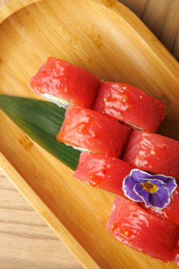 Taze ton balıklı suşi rulosu ahşap bir tepside basit bir çiçek süslemesiyle sunulur. Tabak, malzemelerin canlı renklerini ve doğal sunum tarzını vurgular.