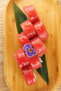 Taze ton balıklı suşi ruloları ahşap bir tabakta düzgünce düzenlenir. Yeşil bir yaprak ve küçük bir dekoratif çiçek sunuma ilave eder. Japon yemeğinden zevk almak için mükemmel..