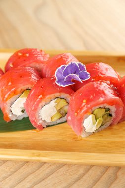 Krem peynir ve sebzelerle dolu suşi ruloları ahşap bir tabakta servis edilir. Parlak kırmızı ton balığı ruloları kaplıyor ve mor bir çiçek süsleme ekliyor..