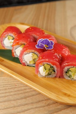 Avokado ve krem peynirle dolu suşi ruloları taze balıkla kaplanır ve çiçekle süslenir. Bu yemek Japon mutfağını temsil ediyor ve canlı tatlar sunuyor..