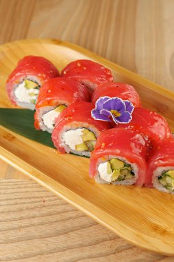 Suşi ruloları ahşap bir tepside canlı kırmızı balıklar, kremalı dolgu ve dekorasyon için küçük mor bir çiçek ile düzenlenir. Yemek taze ve renkli görünüyor..