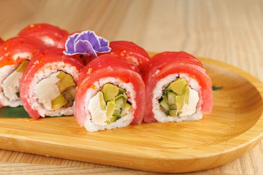 Suşi ruloları taze sebzelerle doludur ve balıklar ahşap bir tabakta çiçek ile süslenir. Bu yemek canlı renkler ve tatlar sergiliyor..