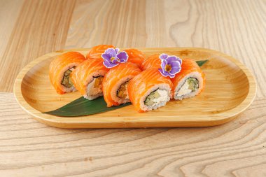 Bir tabakta somon, salatalık ve krem peynirle dolu suşi ruloları var. Ekmekler ahşap bir yüzeyde küçük mor çiçeklerle süslenir..
