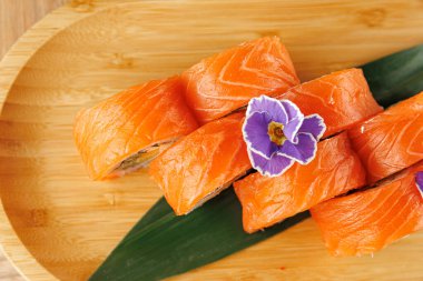 Somonlu taze suşi ruloları ahşap bir tabakta düzenlenir. Mor bir çiçek, kabı süsleyip görünüşünü güçlendirir. Bu sahnede Japon mutfağı sergileniyor..
