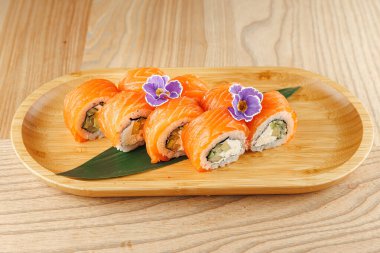 Bambu tabağında somon, avokado ve yenilebilir çiçeklerle süslenmiş salatalık dolu suşi ruloları var. Ayar hafif ahşap bir yüzeye sahip..