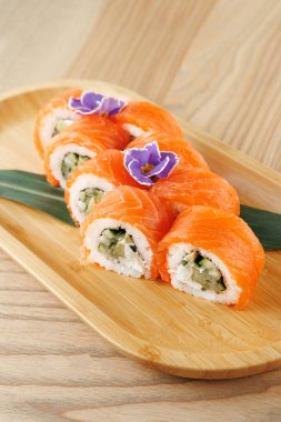 Somon balıklı suşi ruloları ve ahşap bir tabakta düzgünce dizilmiş salatalık. Yenilebilir çiçekler renk katar, yemek ortamında sunumunu güçlendirir..