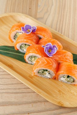 Salatalık ve krem peynirle dolu suşi ruloları ahşap bir tabakta güzelce düzenlenir. Parlak portakallı somon dilimleri ekmeklerin üstünde, süsleme için çiçekler var..