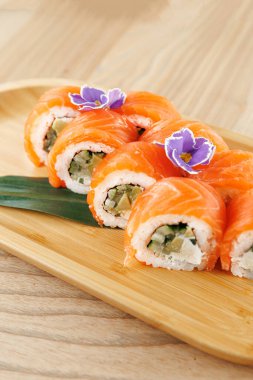 Renkli suşi ruloları ahşap bir tabakta güzelce düzenlenir. Ekmekler balık ve sebze içeriyor ve dekorasyon için yenilebilir çiçeklerle süsleniyorlar. Bu yemek paylaşmak için mükemmel..